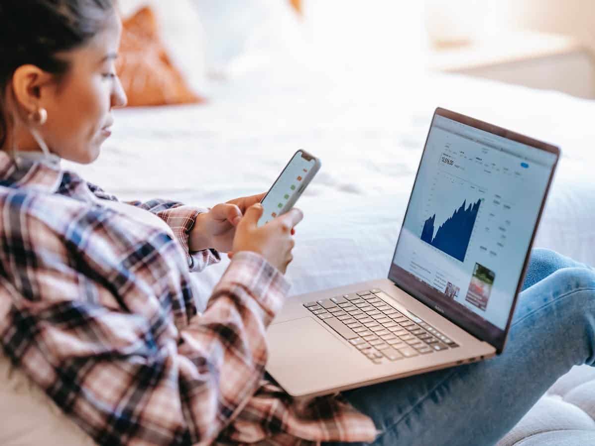 A woman with a laptop on her lap views a graph while also looking at the smart phone she is holding.