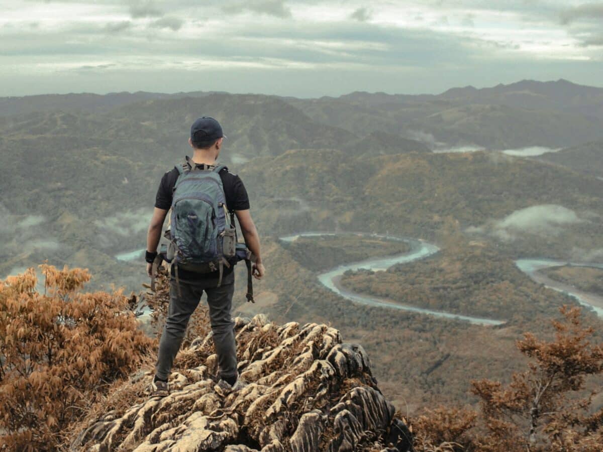 man with backpack stands at the top of a mountain looking out over a river