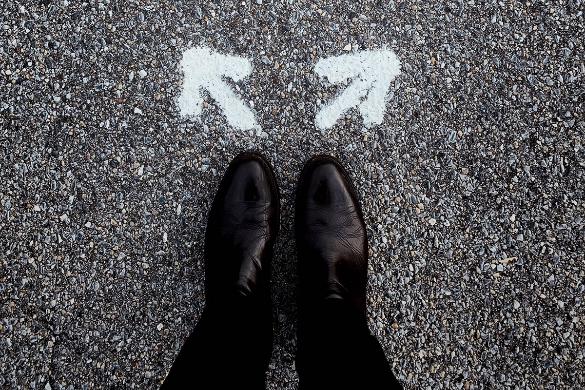 Feet wearing black shoes with white arrows painted on the blacktop just above the toes