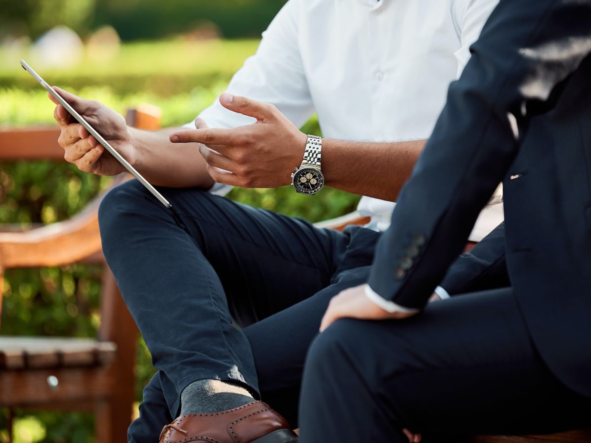 Two men dressed in business attire sit on a park bench. One gestures at the tablet he is holding while the other looks on