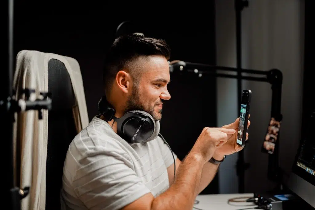 A man is wearing headphones around his neck while recording a vertical video on a smartphone.