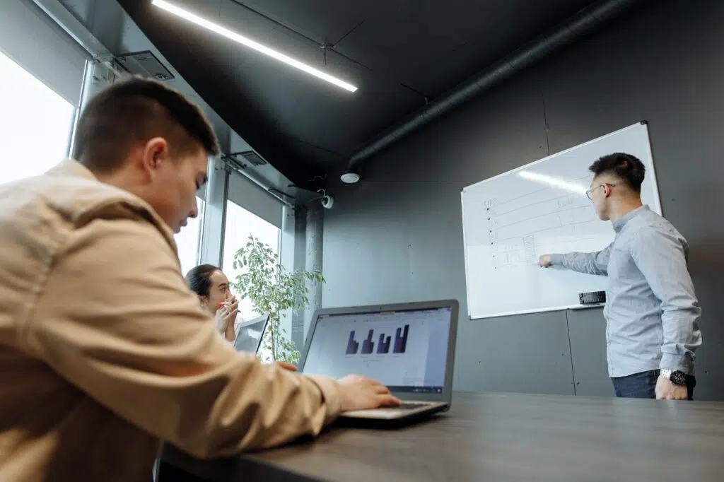 one man stands at a whiteboard while another sits at a laptop. A woman looks on in the background.