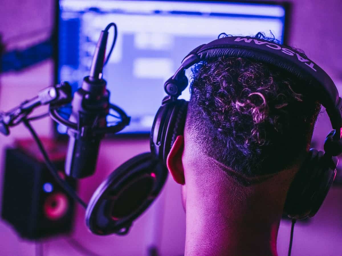 a voiceover artists sits with his back to the camera. he is recording using a microphone, pop filter, and headset. A computer screen is lit up in front of him